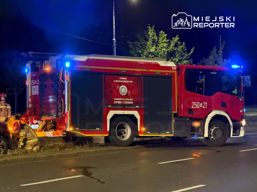 Strażacy w hełmach pracują przy wozie strażackim z włączonymi sygnałami świetlnymi na nocnej ulicy.