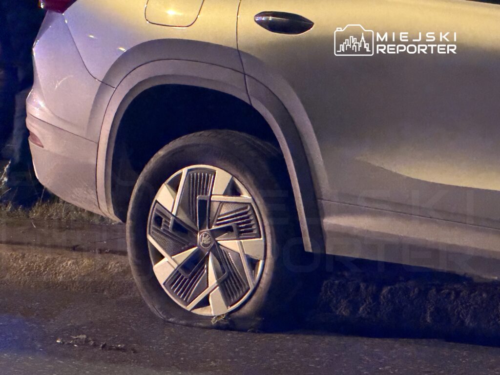 Samochód osobowy z przebitą oponą zaparkowany przy drodze, w tle widoczna trawa i nocne oświetlenie.