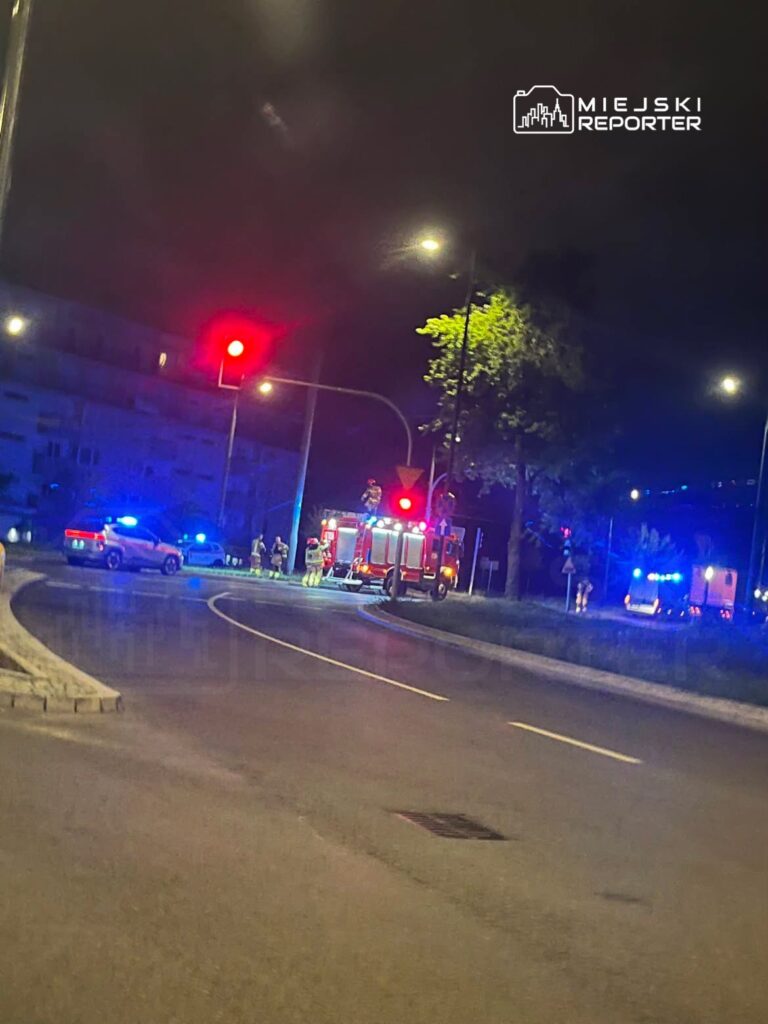 Na skrzyżowaniu w nocy, strażacy i policja interweniują przy włączonych sygnalizacjach świetlnych.
