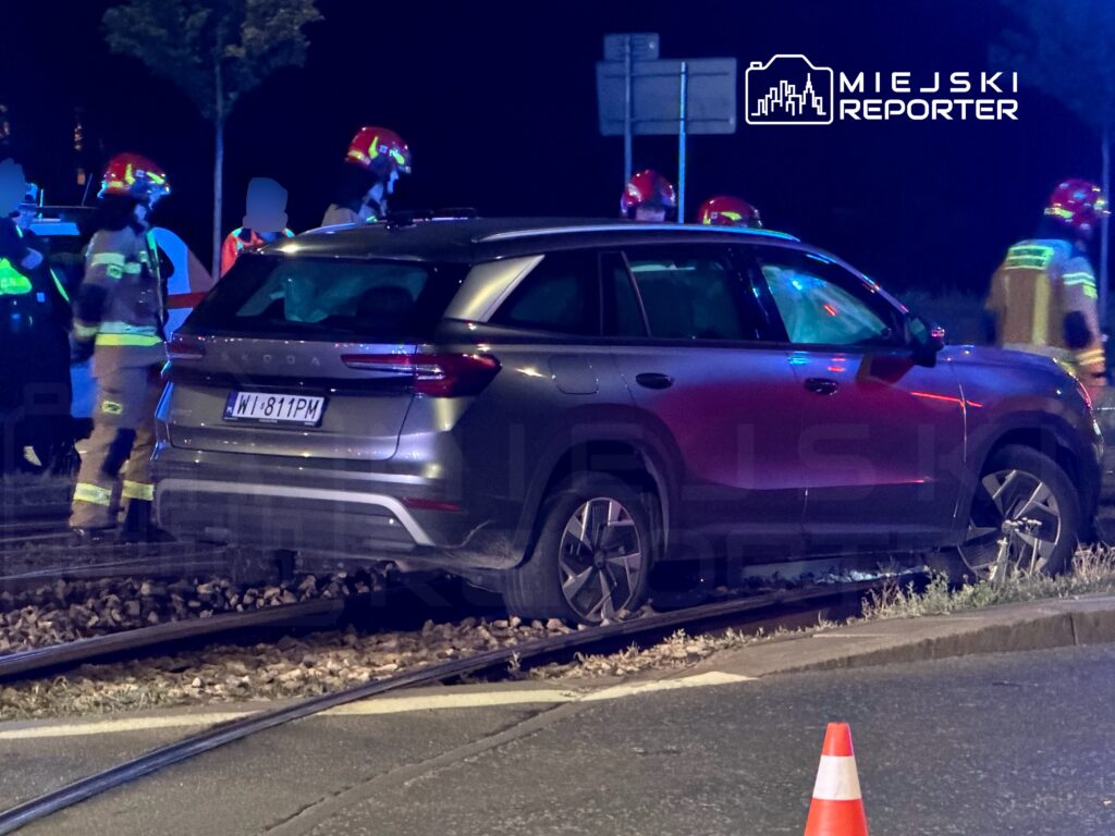 Samochód osobowy stoi na torach kolejowych, obok pracują strażacy w kaskach. W tle widoczne światła sygnalizacyjne.