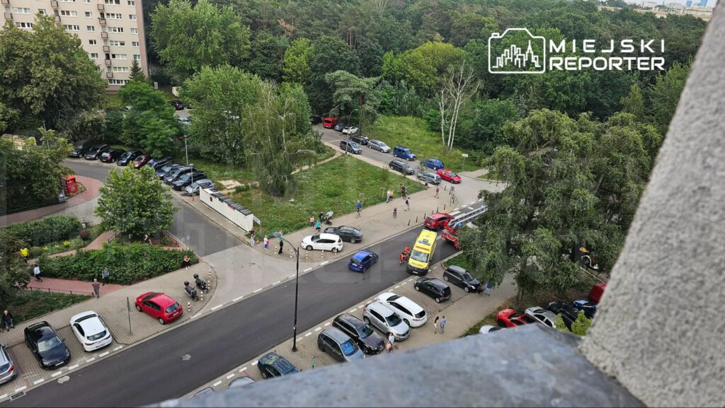 Grupa ludzi obserwująca zdarzenie drogowe w pobliżu nowoczesnych budynków mieszkalnych, z karetką na miejscu.