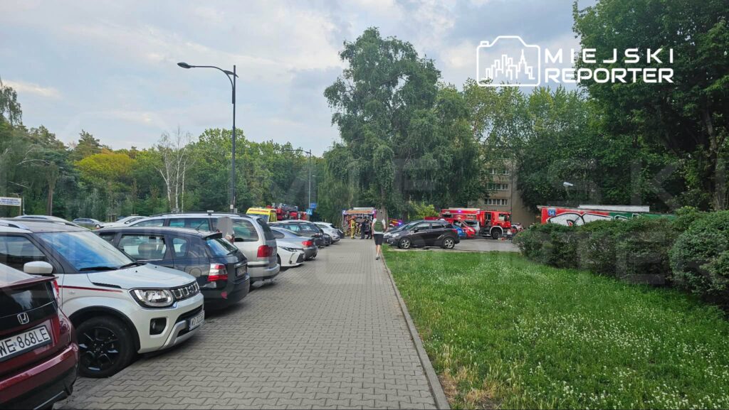 W tle widać pojazdy straży pożarnej oraz ludzi w mundurach, a na pierwszym planie zaparkowane samochody na chodniku.