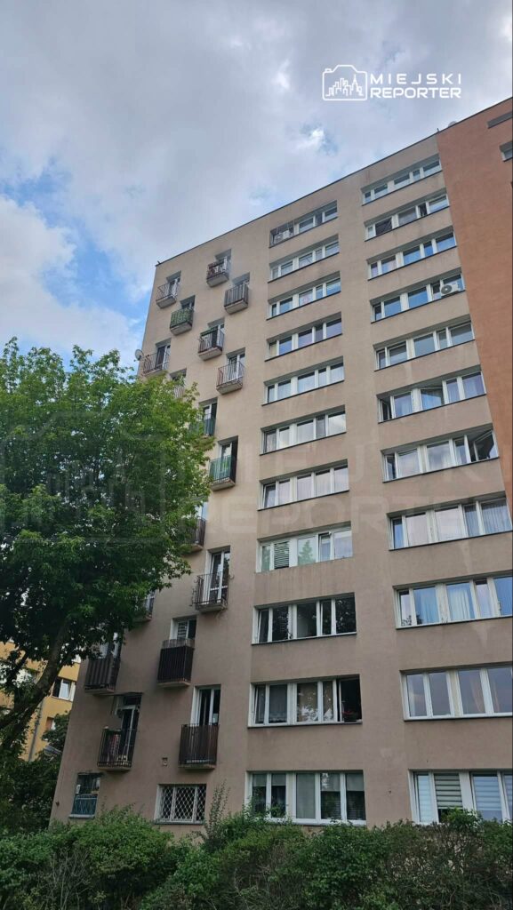 Sześciopiętrowy budynek mieszkalny z balkonami i oknami, otoczony zielenią drzew i krzewów.
