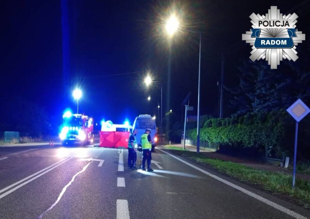 Policyjny radiowóz oraz wóz strażacki na oświetlonej ulicy, gdzie funkcjonariusze rozmawiają obok bariery zabezpieczającej.