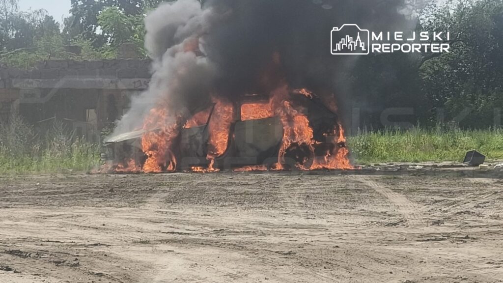 Pojazd w płomieniach, otoczony gęstym dymem, na nieutwardzonej powierzchni w terenie.