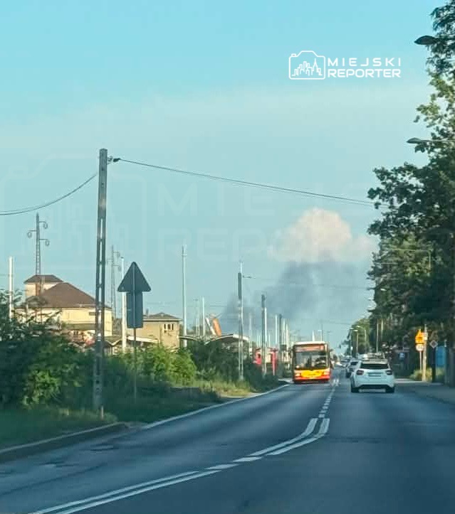 Autobus miejski zatrzymuje się na przystanku przy drodze, w tle widoczny dym unoszący się nad budynkami.