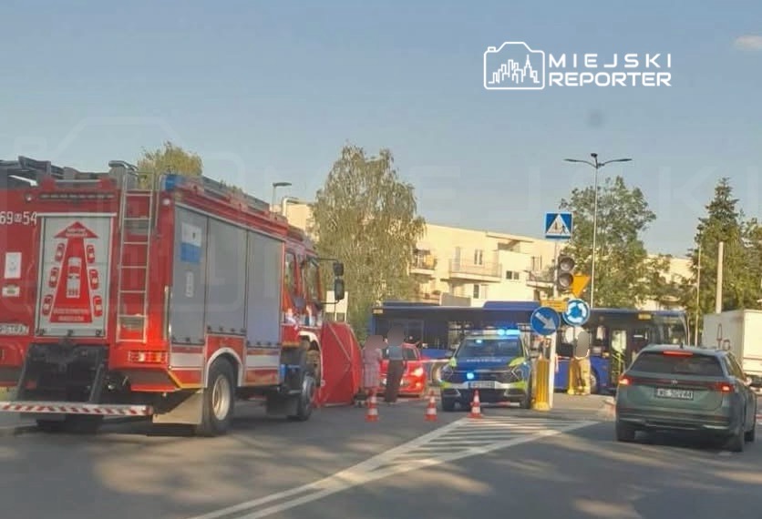 W miejscu zdarzenia znajdują się pojazdy straży pożarnej i policji, a także zablokowany autobus na skrzyżowaniu.