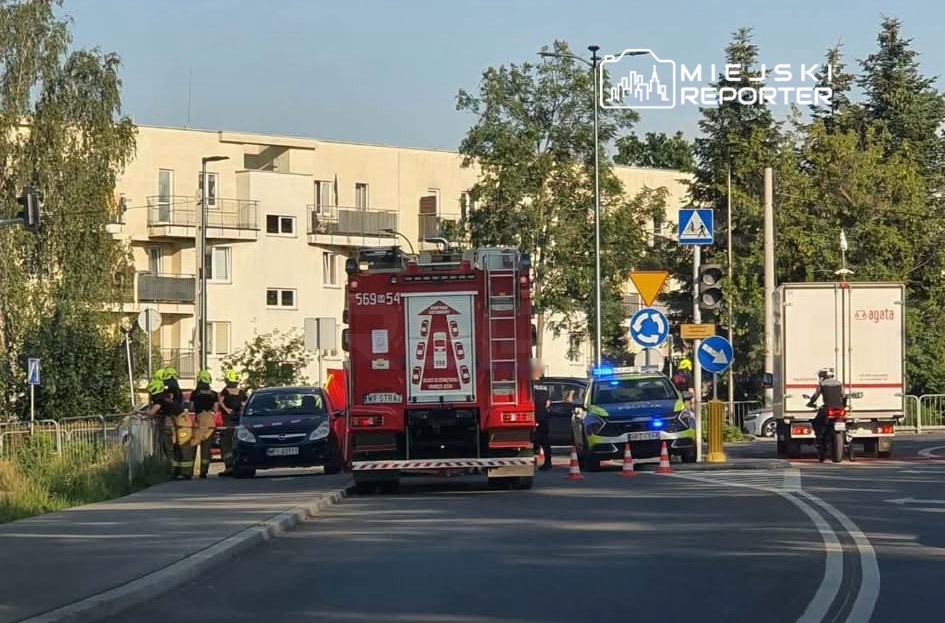 Grupa strażaków i policjantów zabezpiecza miejsce zdarzenia obok czerwonego wozu strażackiego na miejskiej ulicy.