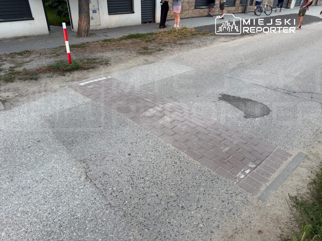 Na ulicy widoczny jest nowo wykonany fragment chodnika z kostki brukowej, otoczony szaro-skalistą nawierzchnią. W tle stoją przechodnie oraz rowerzysta.