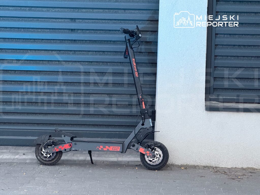 Skuter elektryczny zaparkowany przy szarej ścianie budynku, obok metalowych żaluzji.