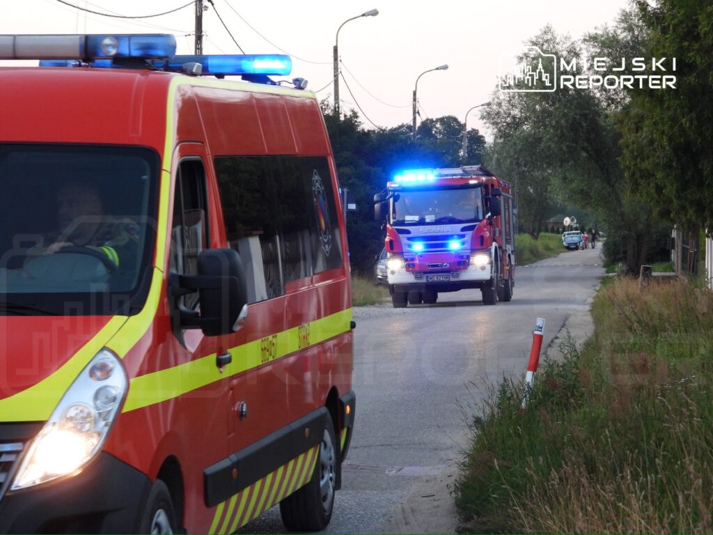 Czerwony samochód strażacki z włączonymi sygnalizatorami jedzie wzdłuż wiejskiej drogi, za nim ambulans.