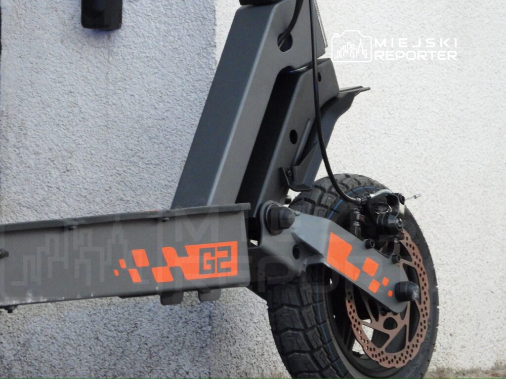 Skuter elektryczny z widocznym logo "G2" oparty na szarej ścianie, z wyraźnie widoczną tylną oponą i hamulcem tarczowym.