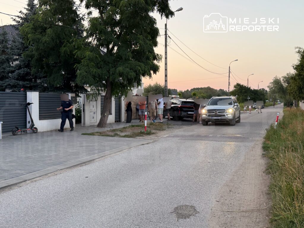 Grupa osób rozmawia na chodniku obok zaparkowanego samochodu, a policjant prowadzi rozmowę z jednym z uczestników.