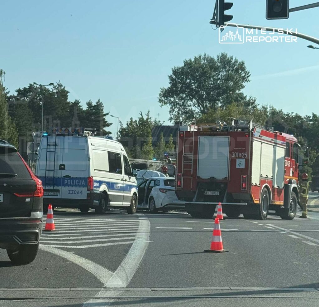 Na skrzyżowaniu zatrzymane pojazdy, w tym radiowóz i wóz strażacki, oraz służby ratunkowe zabezpieczające miejsce zdarzenia.