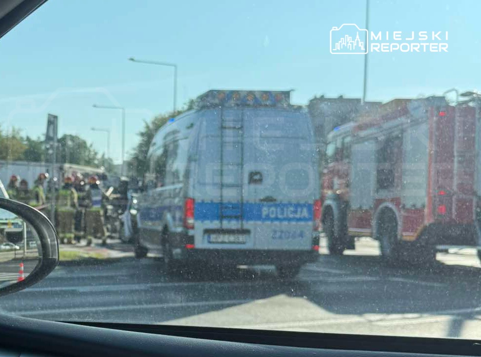 Grupa strażaków i policjantów pracuje przy wypadku drogowym, obok zaparkowanych pojazdów ratunkowych.