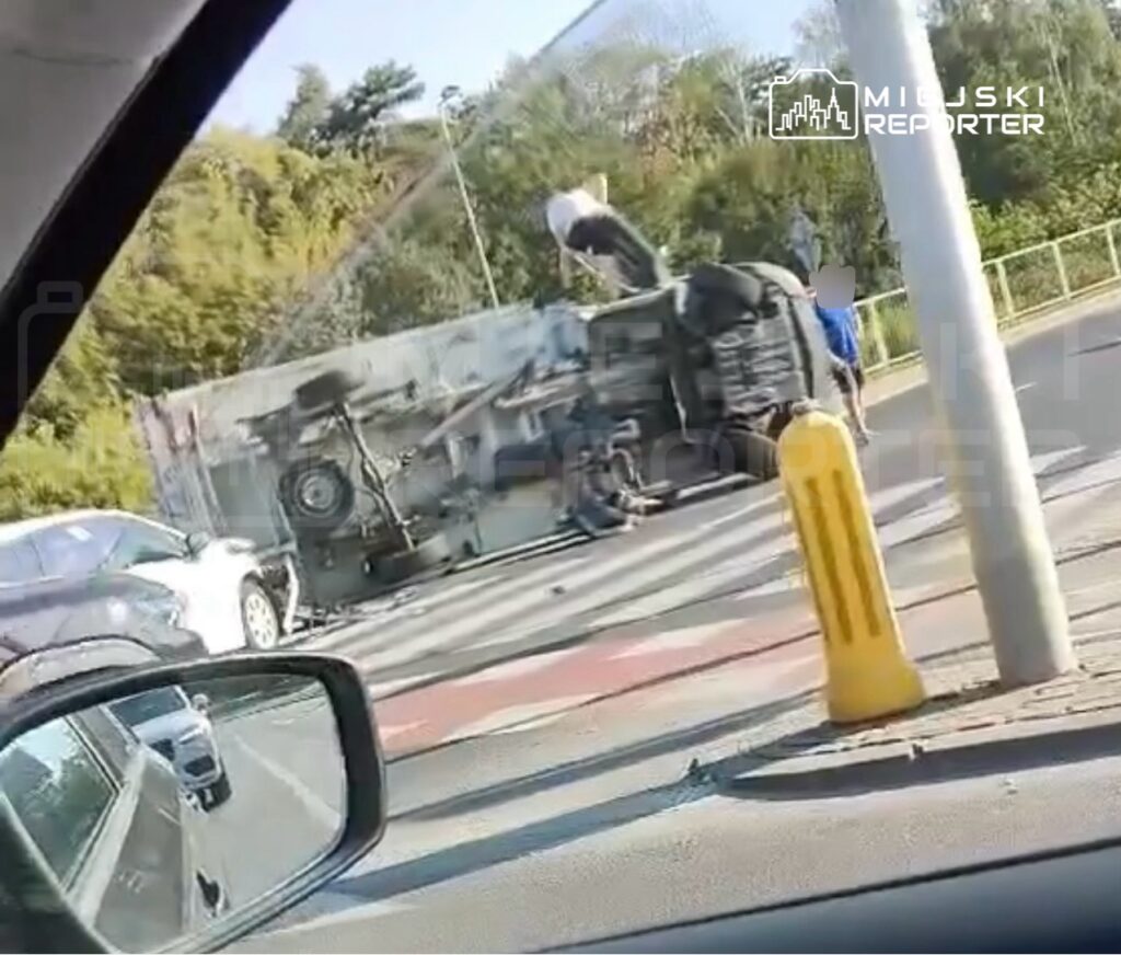 Na drodze leży przewrócone auto, a obok grupka osób próbuje pomóc poszkodowanym. W tle widać inne pojazdy.