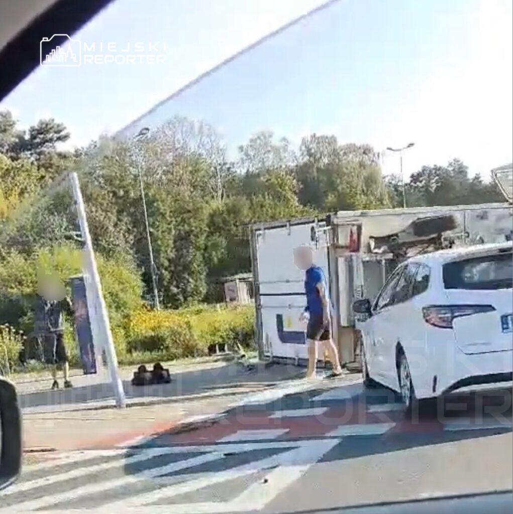 Mężczyzna w niebieskiej koszulce przechodzi obok przewróconego kontenera na drodze, obok białego samochodu.