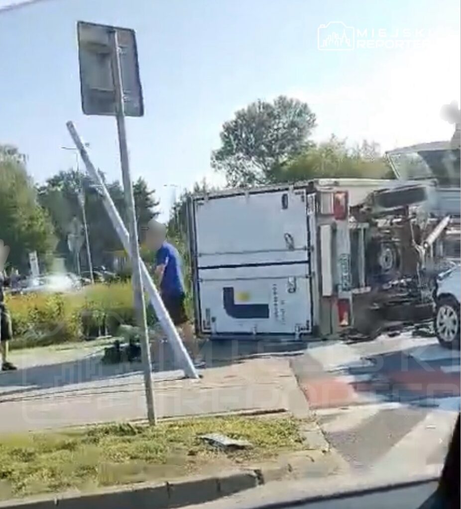 Przewrócony pojazd dostawczy na drodze, obok mężczyzna w niebieskiej koszulce, w tle uszkodzone auto i latarnia.