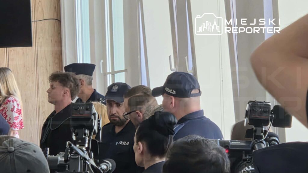 Grupa policjantów w mundurach stoi w pomieszczeniu z drewnianymi ścianami, otoczona przez dziennikarzy z kamerami.
