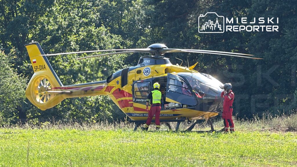 Śmigłowiec ratunkowy w żółto-czerwonym malowaniu lądował na trawie, a ratownicy w kombinezonach przygotowują się do akcji.