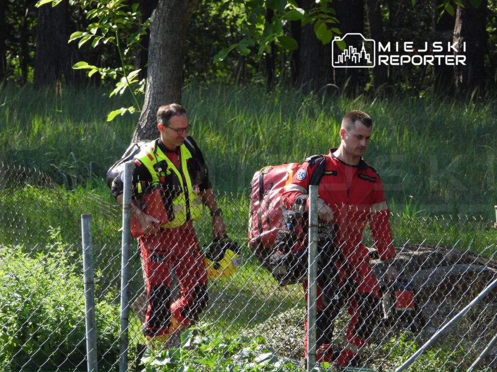 Dwaj ratownicy w odzieży ochronnej przechodzą przez ogrodzenie w otoczeniu zieleni, niosąc sprzęt ratunkowy.