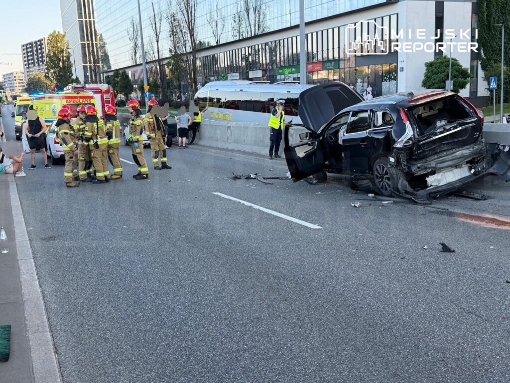 Grupa strażaków w mundurach bada miejsce wypadku drogowego obok uszkodzonego samochodu osobowego.
