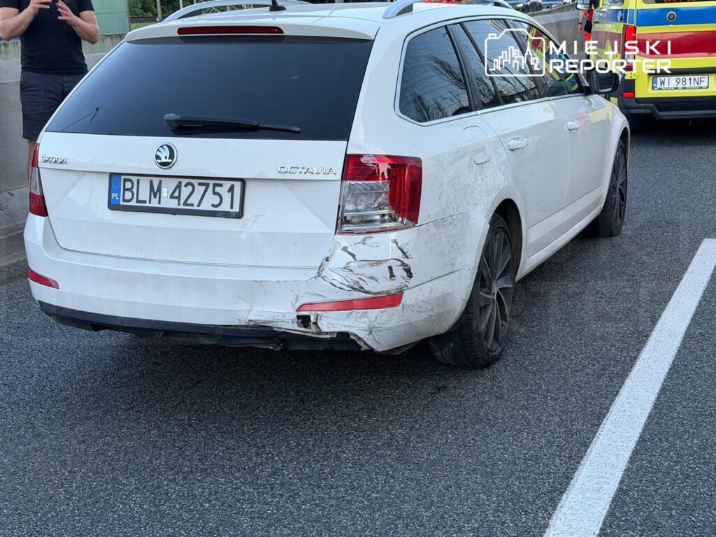 Biały samochód osobowy marki Skoda Octavia z uszkodzeniem tyłu stoi na jezdni, obok mężczyzna rozmawia przez telefon.