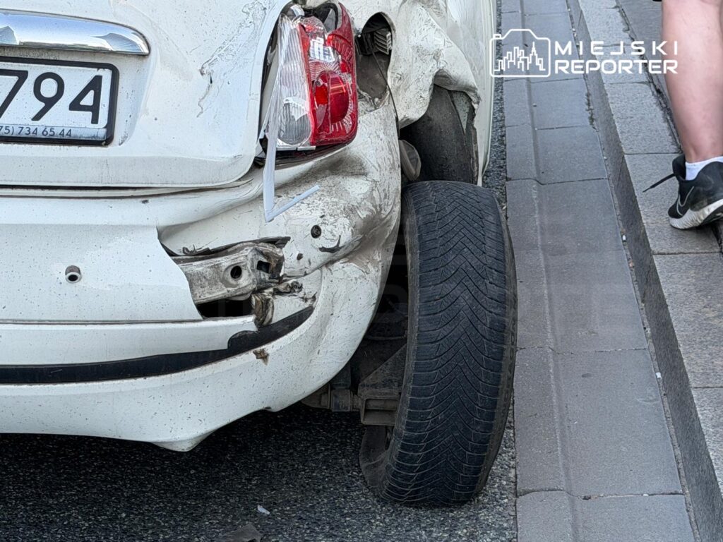 Biały samochód osobowy z uszkodzoną tylną częścią stoi na chodniku obok zniszczonej opony.