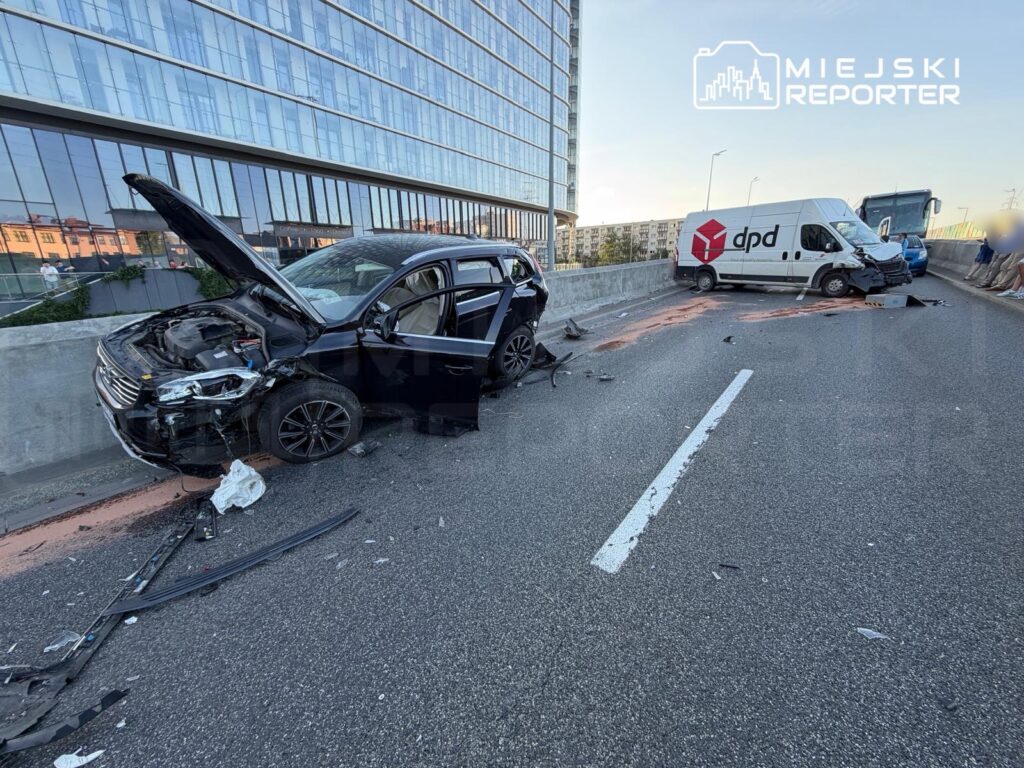 Zniszczony czarny samochód osobowy z otwartą maską stoi na jezdni obok białego vana z logo DPD, w otoczeniu nowoczesnych budynków.