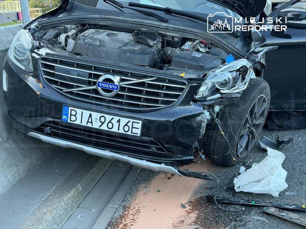 Uszkodzony czarny samochód marki Volvo z otwartą maską stoi na poboczu drogi, obok leżą fragmenty zderzaka.
