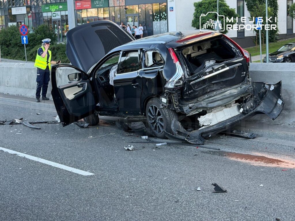 Rozbity samochód osobowy z otwartymi drzwiami stoi na jezdni, obok policjant w kamizelce odblaskowej prowadzi czynności.