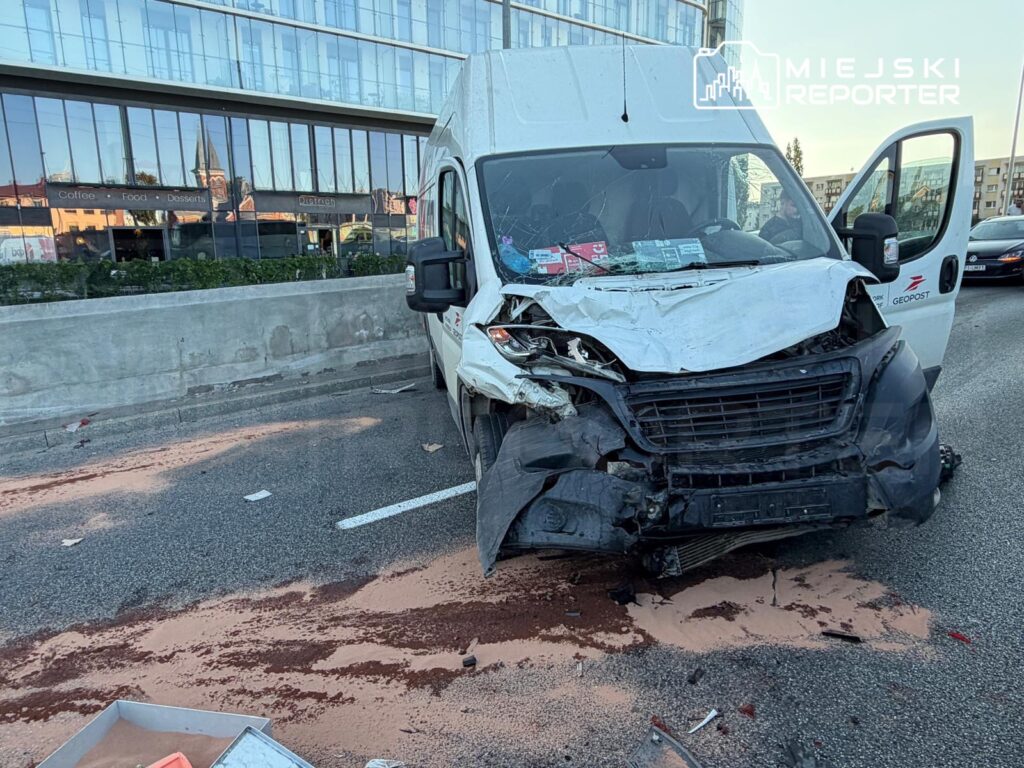 Rozbity samochód dostawczy z uszkodzoną przednią częścią stoi na jezdni, obok nowoczesnego budynku z przeszkloną fasadą. Na drodze widoczne ślady po rozlanych substancjach.