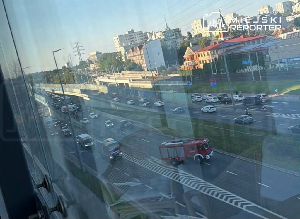 Ruch samochodowy na drodze z widokiem na zatrzymane pojazdy oraz wjeżdżającą straż pożarną.