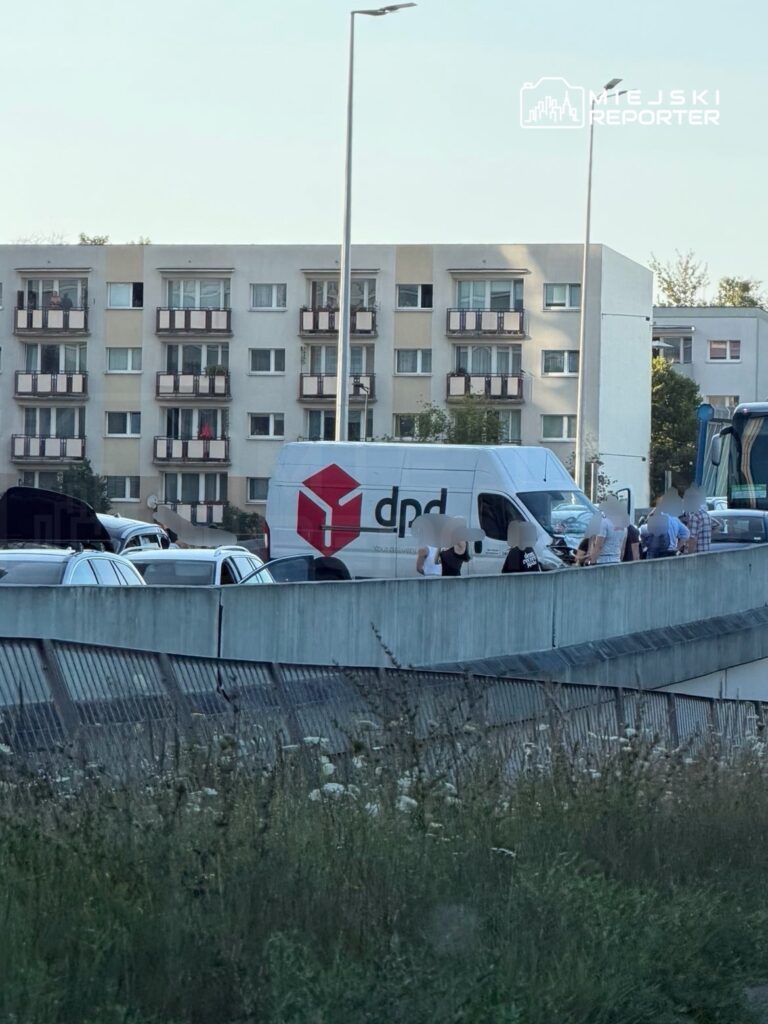 Kierowca dostawczego samochodu DPD rozmawia z grupą ludzi na parkingu przy budynkach mieszkalnych.