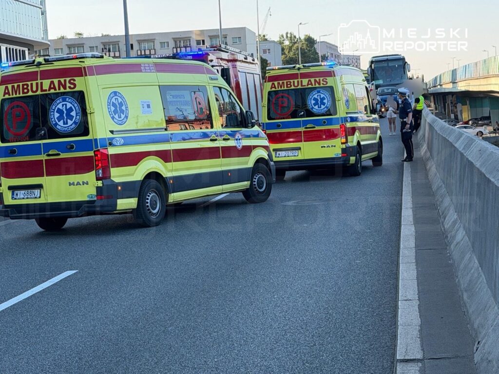 Dwa karetki pogotowia z włączonymi sygnałami świetlnymi zatrzymane na jezdni, obok policjant rozmawiający z przechodniami.