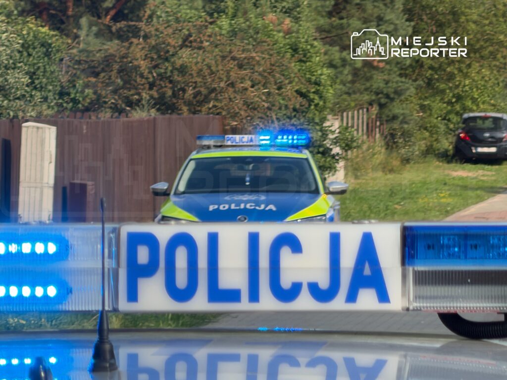 Nieoznakowany radiowóz policyjny z włączonymi sygnałami świetlnymi stojący na drodze w otoczeniu zieleni.