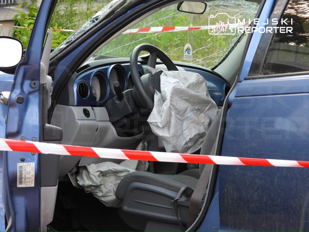 Wnętrze niebieskiego samochodu z wystrzelonymi poduszkami powietrznymi, otoczone taśmą policyjną.