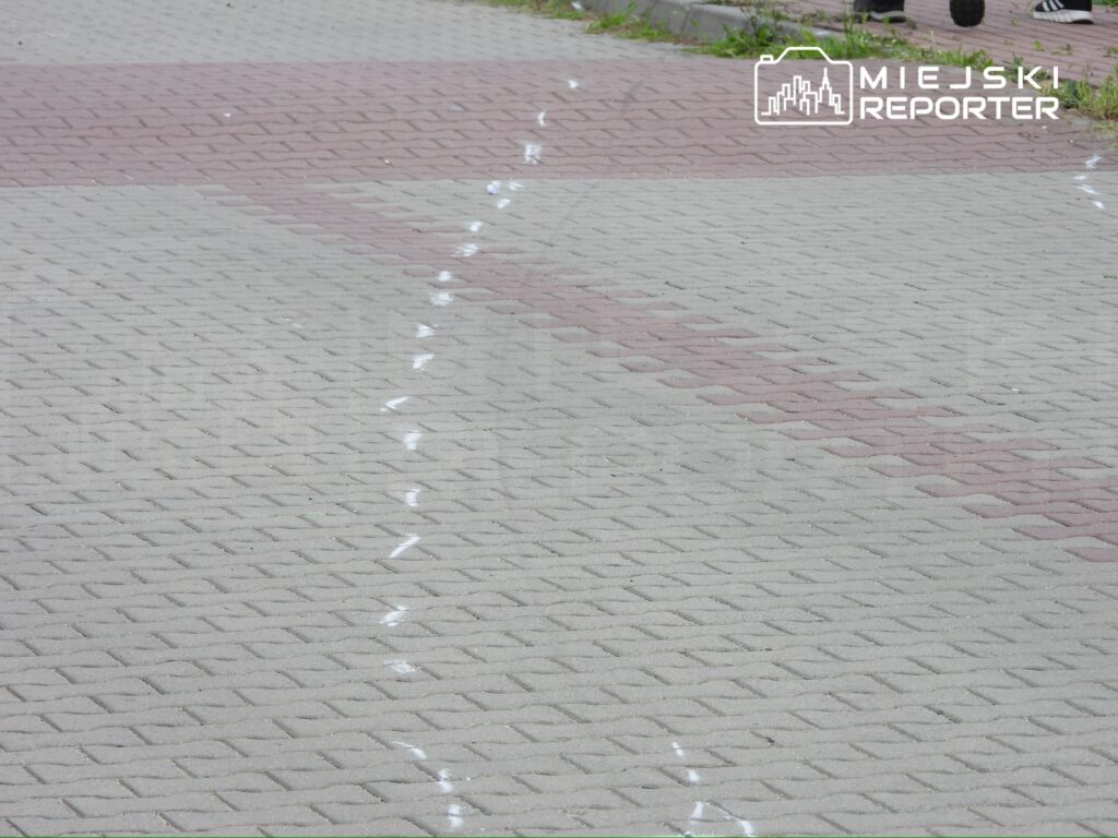 Biała linia na chodniku prowadząca do miejsca zdarzenia, otoczona szaro-beżowymi płytkami.
