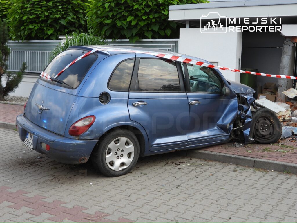 Nieoznakowany niebieski samochód osobowy uderzył w ścianę budynku, z widocznymi uszkodzeniami przodu, w otoczeniu zieleni.