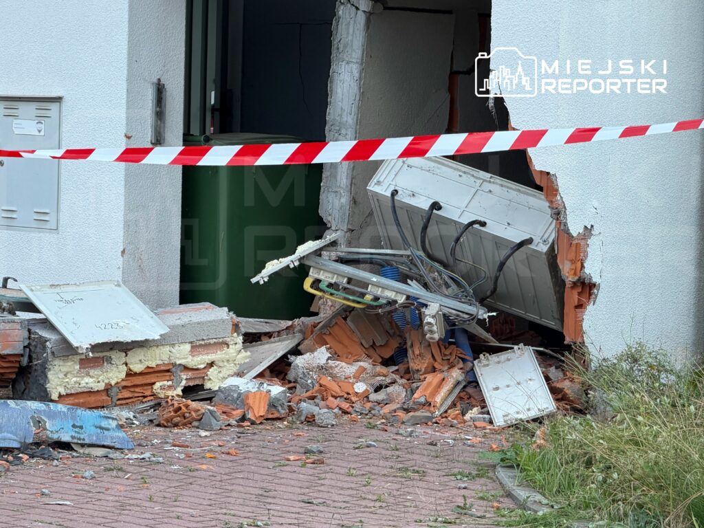 Zniszczona ściana budynku z odsłoniętymi cegłami i gruzem, obok leży uszkodzona skrzynka oraz zabezpieczenie w postaci taśmy.