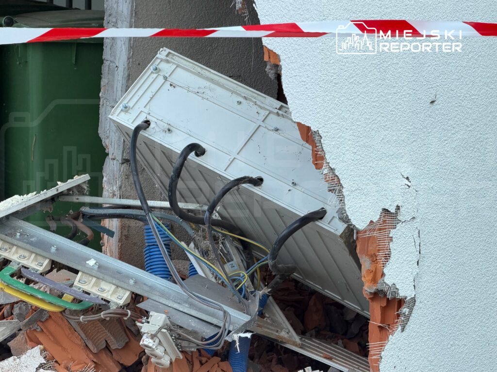 Uszkodzony fragment ściany budynku z wystającymi przewodami i elementami instalacji, otoczony taśmą ostrzegawczą.