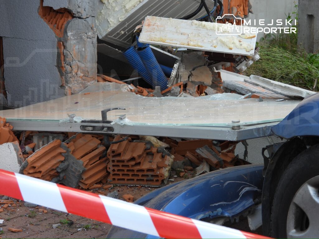 Zniszczona ściana budynku z odsłoniętymi cegłami i gruzem, obok leży szklana płyta, otoczona taśmą ostrzegawczą.