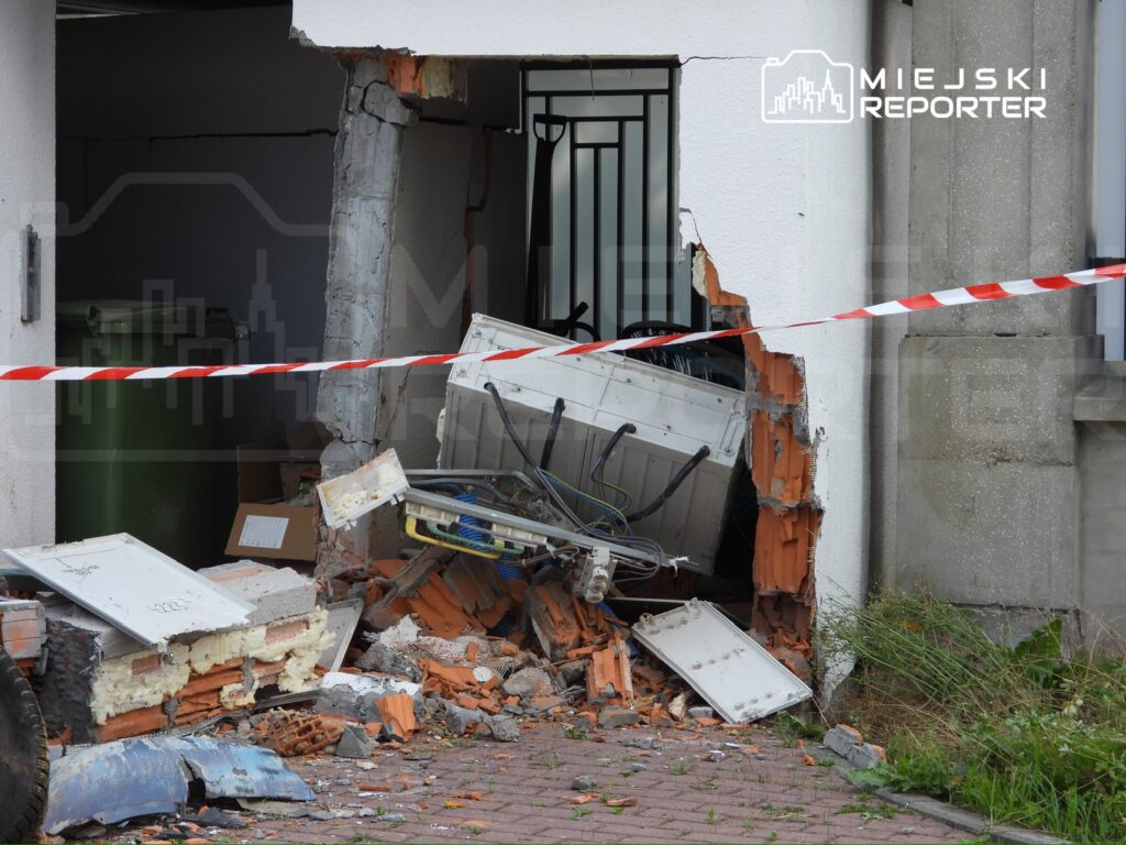 Uszkodzona ściana budynku z widocznym zniszczeniem, w tle sprzęt elektroniczny i materiały budowlane, otoczone taśmą ostrzegawczą.