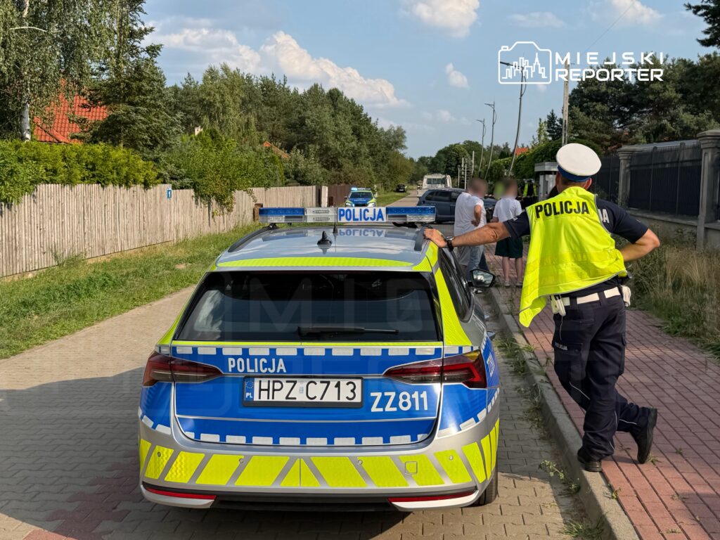 Policjant w kamizelce odblaskowej rozmawia z grupą osób obok radiowozu na osiedlowej drodze.