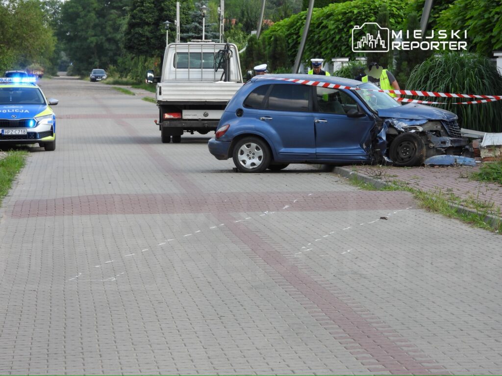 Na drodze stoi niebieski samochód osobowy z uszkodzoną przednią częścią, obok policjanci zabezpieczają miejsce zdarzenia.