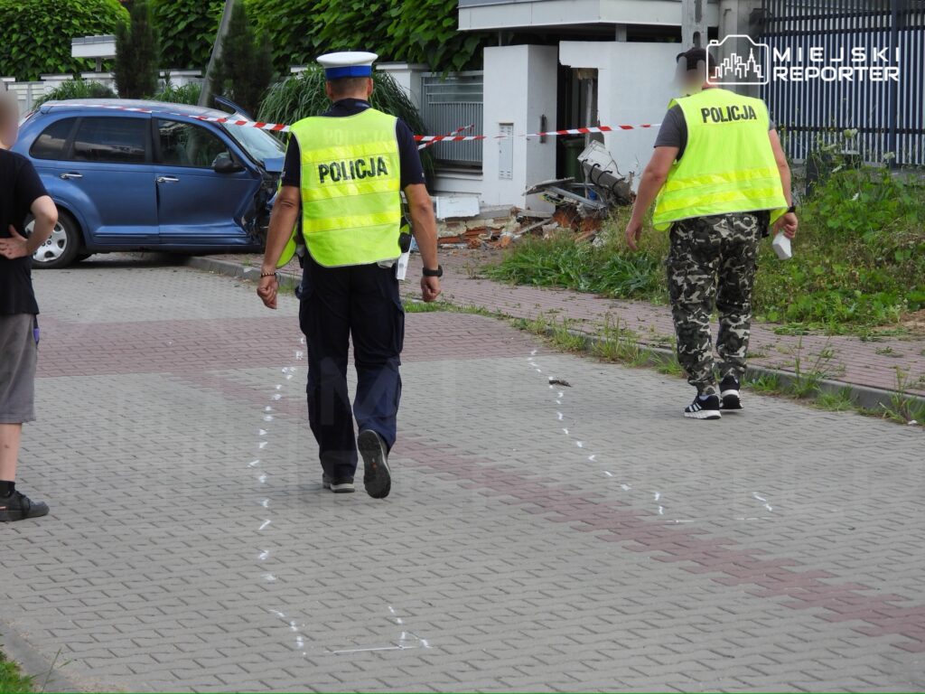 Dwaj policjanci w kamizelkach odblaskowych badają miejsce zdarzenia na chodniku obok uszkodzonego samochodu.