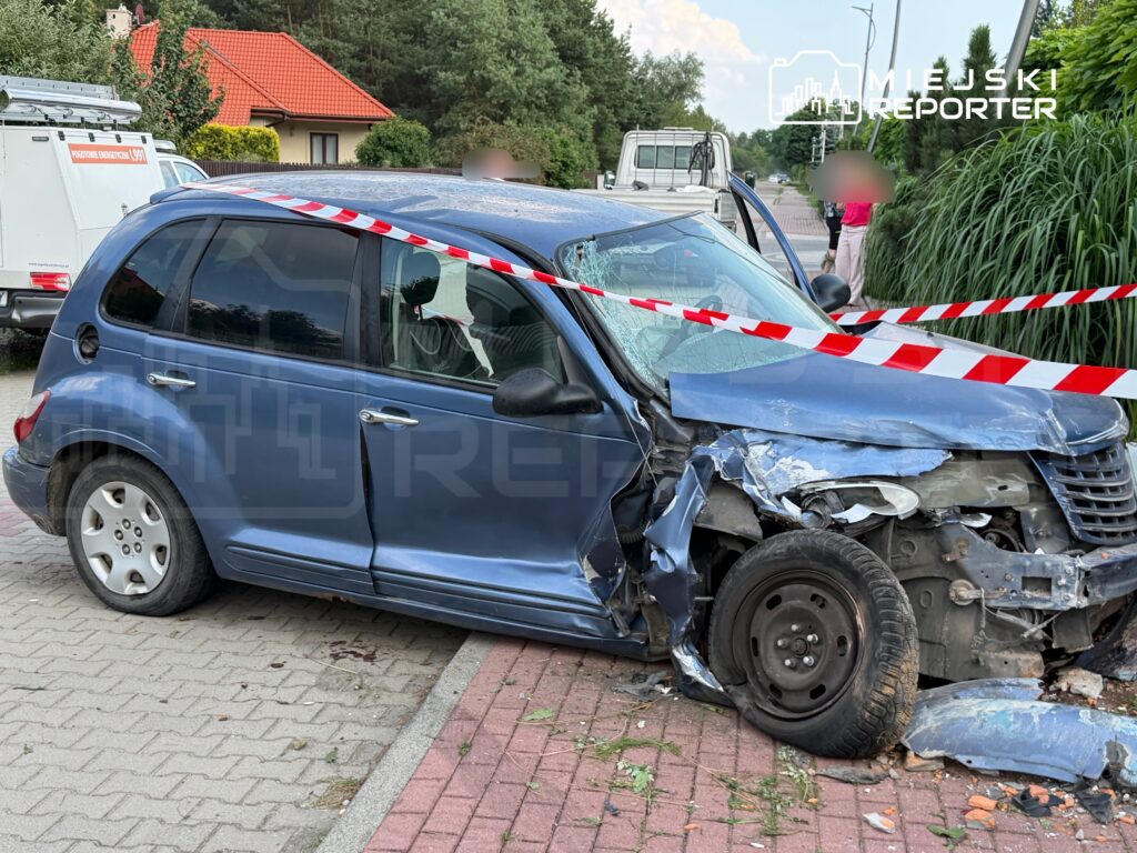 Uszkodzony niebieski samochód osobowy stoi na chodniku, otoczony taśmą ostrzegawczą. W tle widać zieleń oraz budynki mieszkalne.