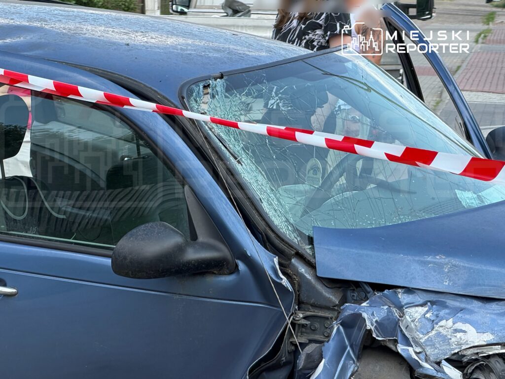 Zniszczony niebieski samochód osobowy z rozbitym przednim szybą, ogrodzony taśmą ostrzegawczą.