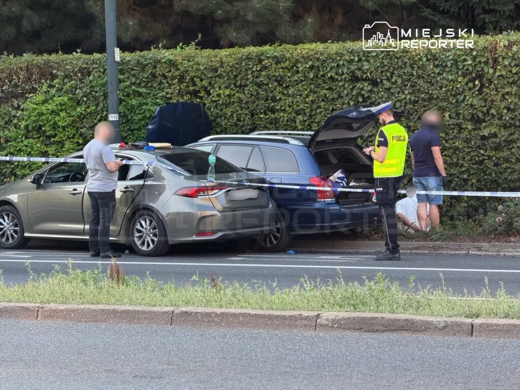 Mężczyzna w szarym t-shircie sprawdza uszkodzenia samochodu, obok policjant w kamizelce odblaskowej notuje informacje przy otwartym bagażniku.