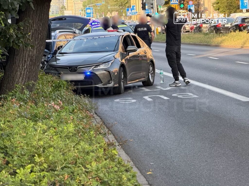 Mężczyzna w czarnym dresie z uniesionymi rękami stoi obok zatrzymanego samochodu osobowego, na drodze w mieście.